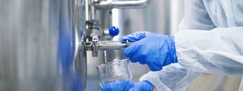 Close up view of hands in protective gloves opening tap valve and filling glass with samples from industrial machine reservoir.